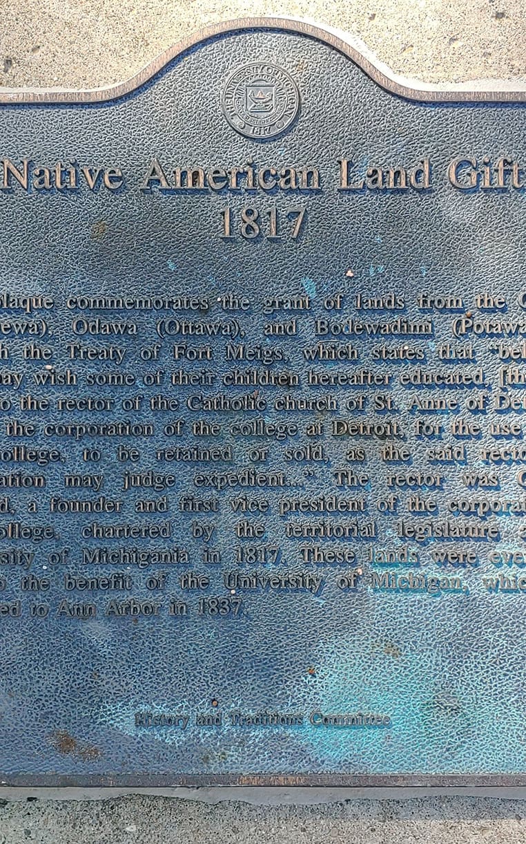 Plaque text reads "Native American Land Gift 1817. This plaque commemorates the grant of lands from the Ojibwe (Chippewa), Odawa (Ottawa), and Bodewadimi (Potawatomi), through the Treaty of Fort Meigs, which states that “believing they may wish some of their children hereafter educated , [they] do grant to the rector of the the Catholic church of St. Anne of Detroit … and to the corporation of the college at Detroit, for the use of the said college, to be retained or sold, as the said rrector and corporation may judge expedient … ” The rector was Gabriel Richard, a founder and first vice president of the corporation of the college, chartered by the territorial legislature as the University of Michigania in 1817. These lands were eventually sold to the beneift of the University of Michigan, which was relocated to Ann Arbor in 1837."