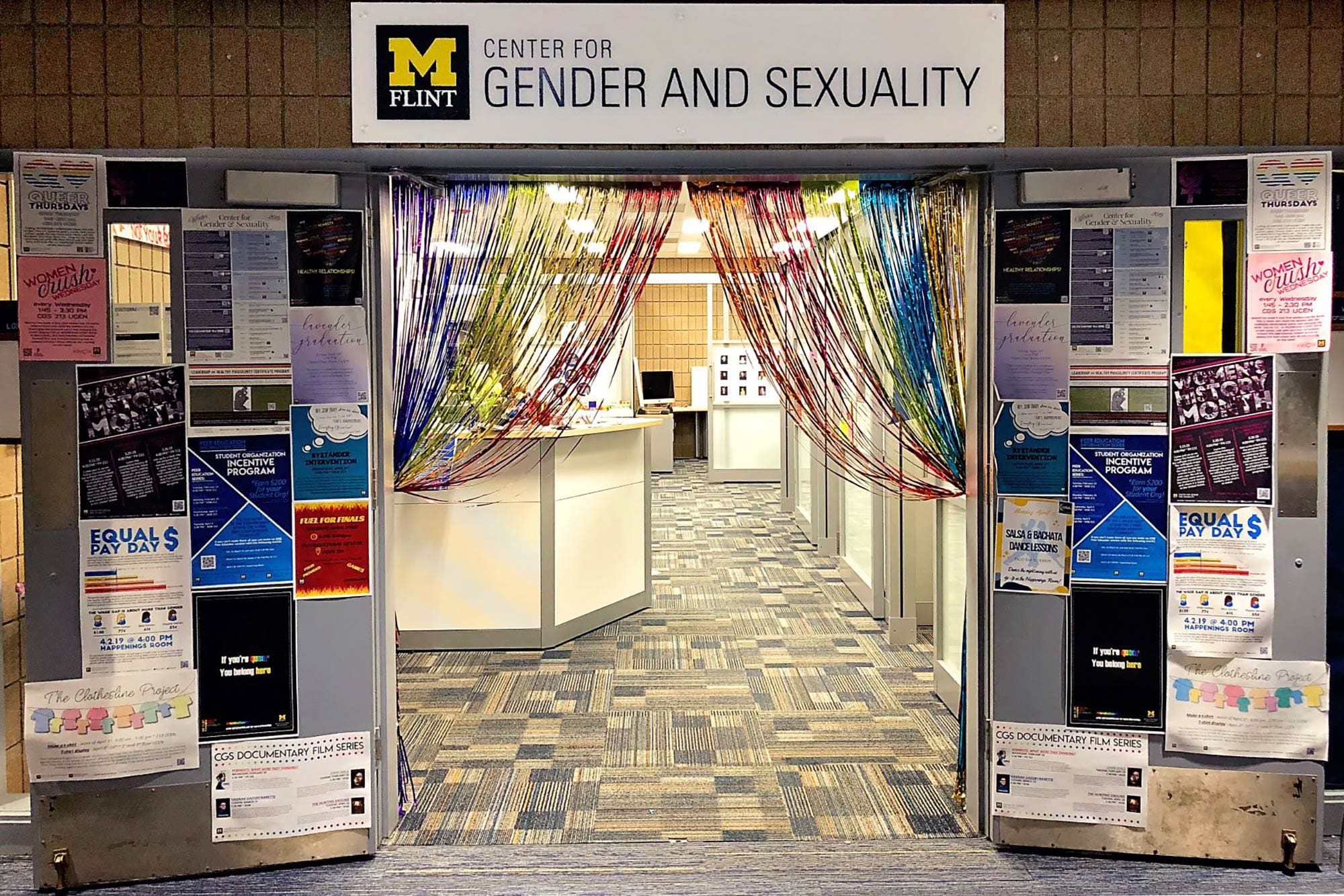 The entrance to UM-Flint's Center for Gender and Sexuality, with two doors open and covered with a variety of posters and announcements, rainbow streamers hanging in the doorframe, and a view into the center itself.