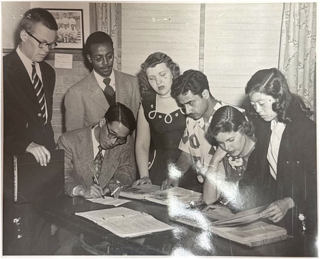 Group of people sitting ad standing around a table, looking at books.
