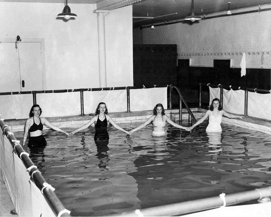 Four women stand in small indoor swimming pool, touching hands to show width of the small pool.
