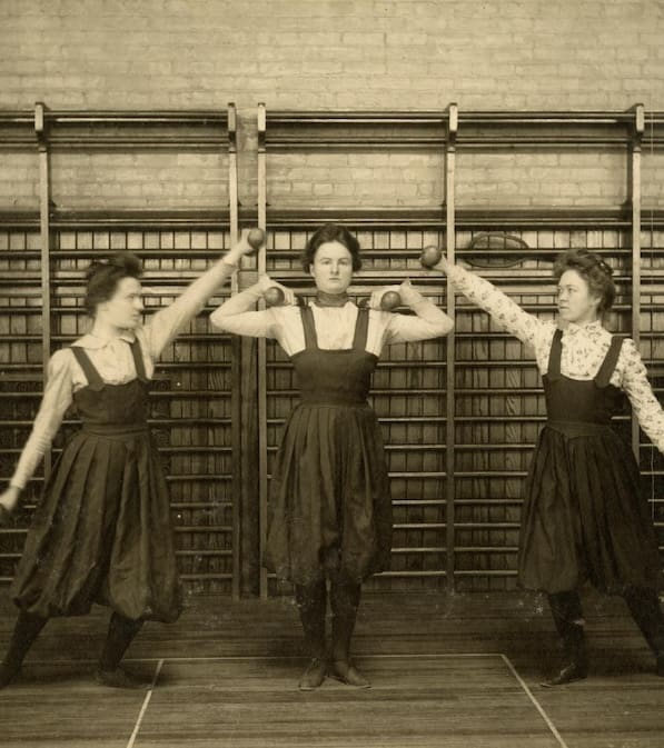 Women in Edwardian pinafore dresses lift dumbells.