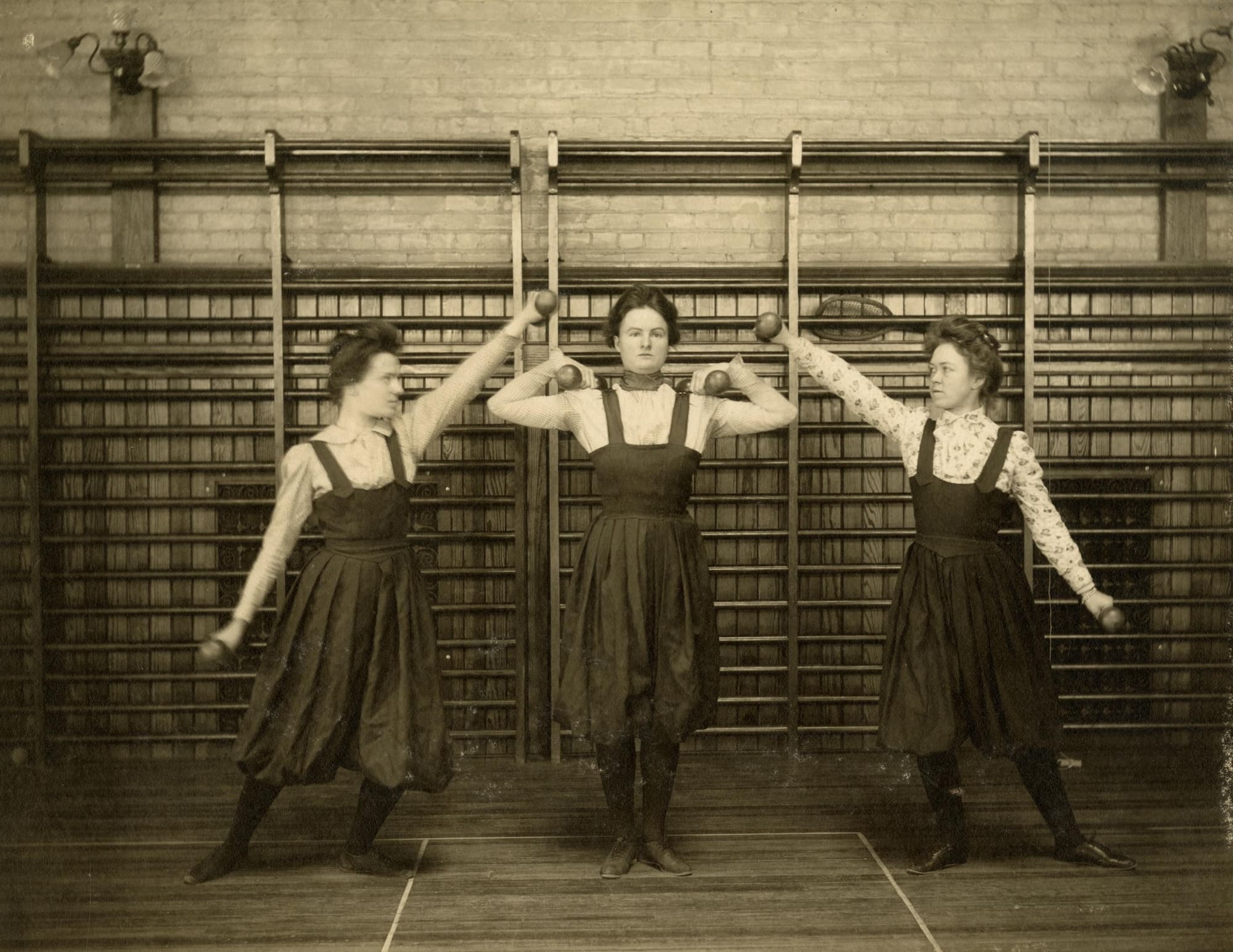 Women in Edwardian pinafore dresses lift dumbells.