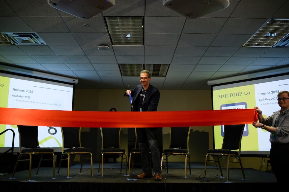 Man stands on stage and cuts a red ribbon with large scissors.