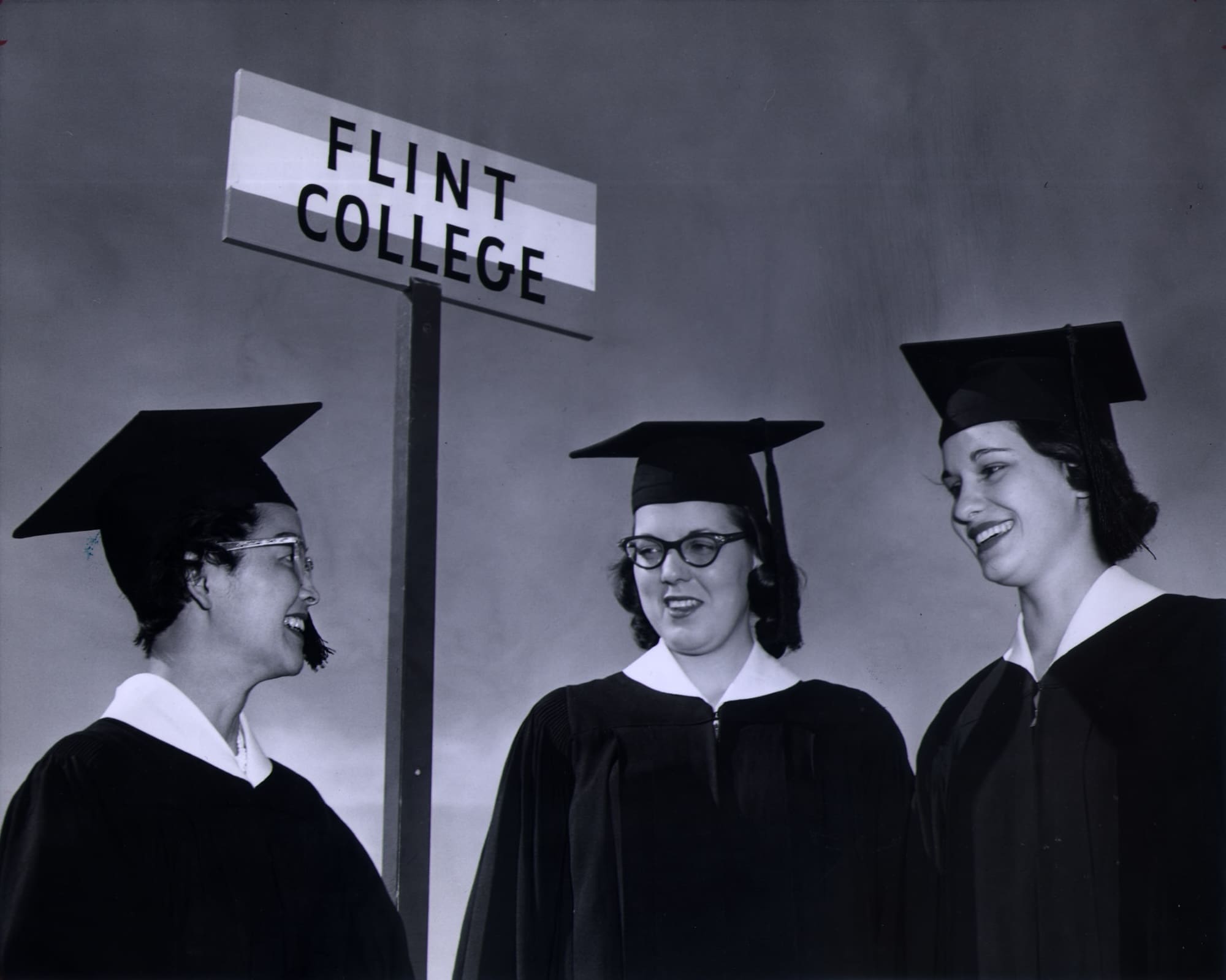 Three women in graduation caps and gowns talk to each other with the "Flint College" sign above their heads in the background.
