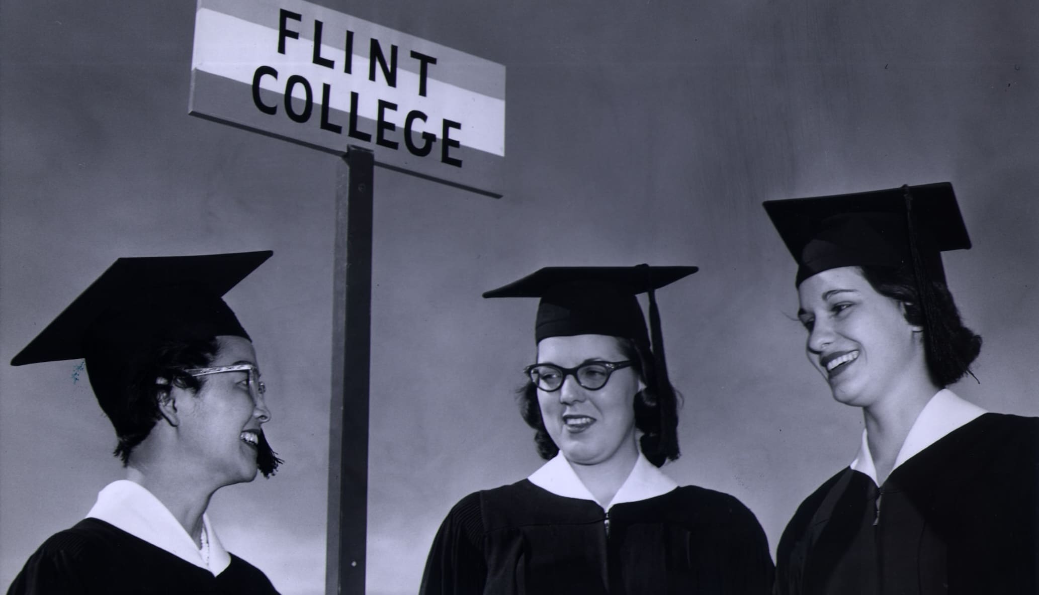 Three women in graduation caps and gowns talk to each other with the "Flint College" sign above their heads in the background.