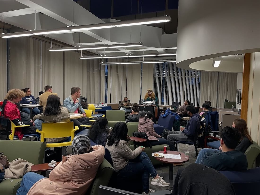 Group of students sit at tables, watching as a fellow student performs at the front of the room.