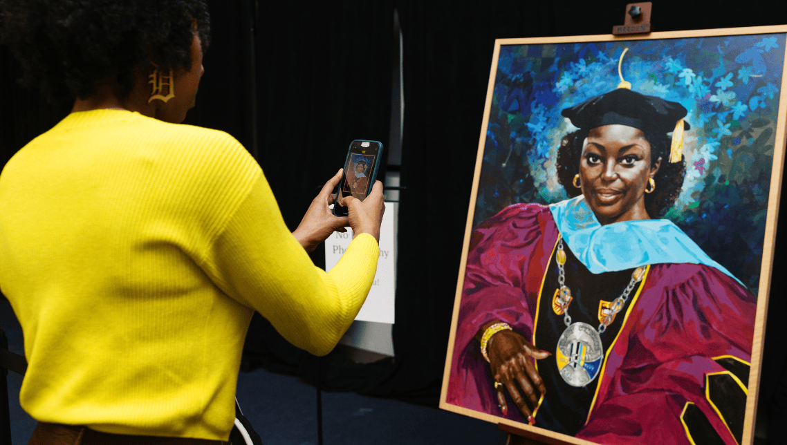 A person takes a smartphone photo of a large painted portrait on an easel depicting Chancellor Blenda Wilson in regalia.