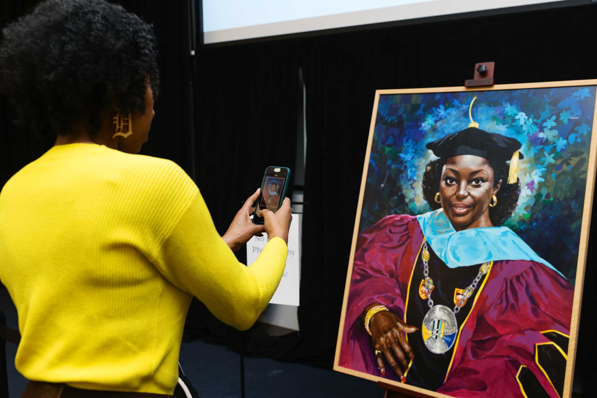 A person takes a smartphone photo of a large painted portrait on an easel depicting Chancellor Blenda Wilson in regalia.