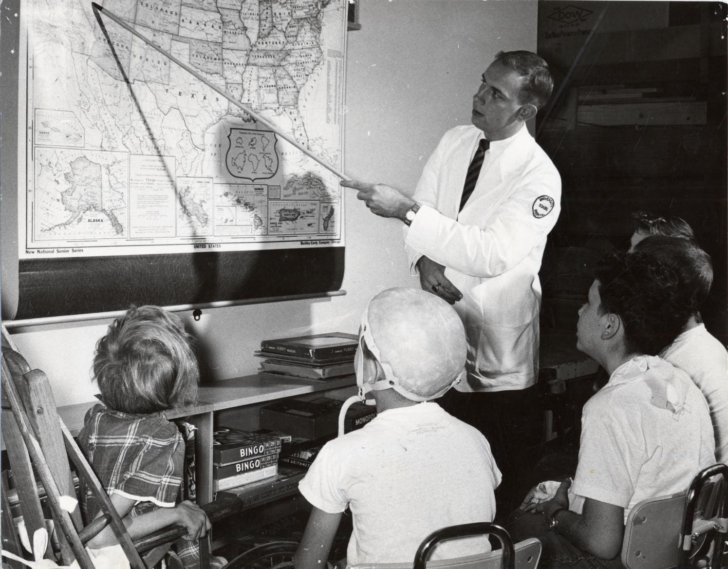 Teacher in white coat points at a map on a classroom board as children from the University Hospital School look at the map.