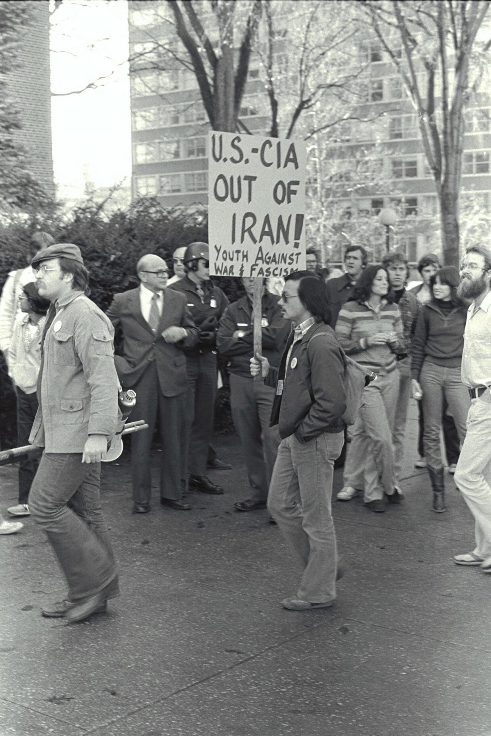 Young man walks through a crowd during a protest on November 20, 1979, he is carrying a picket sign that reads "U.S.-CIA Out of Iran! Youth against war and fascism".