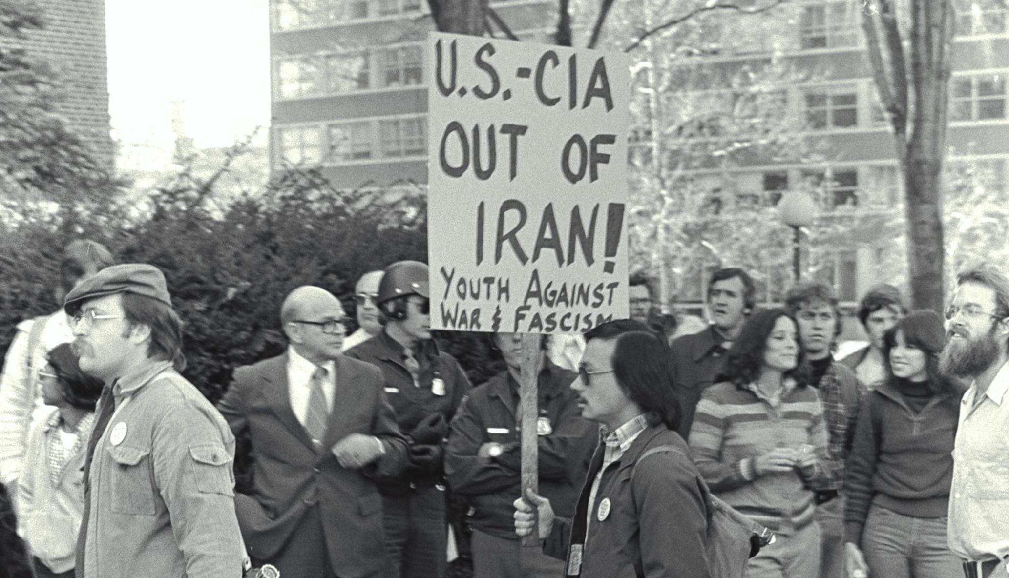 Young man walks through a crowd during a protest on November 20, 1979, he is carrying a picket sign that reads "U.S.-CIA Out of Iran! Youth against war and fascism".